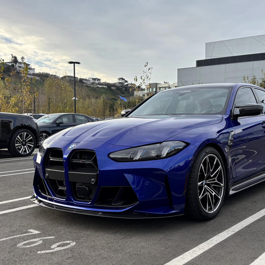 A blue BMW G8X M3/M4 with a carbon fiber front lip installed on the front bumper.