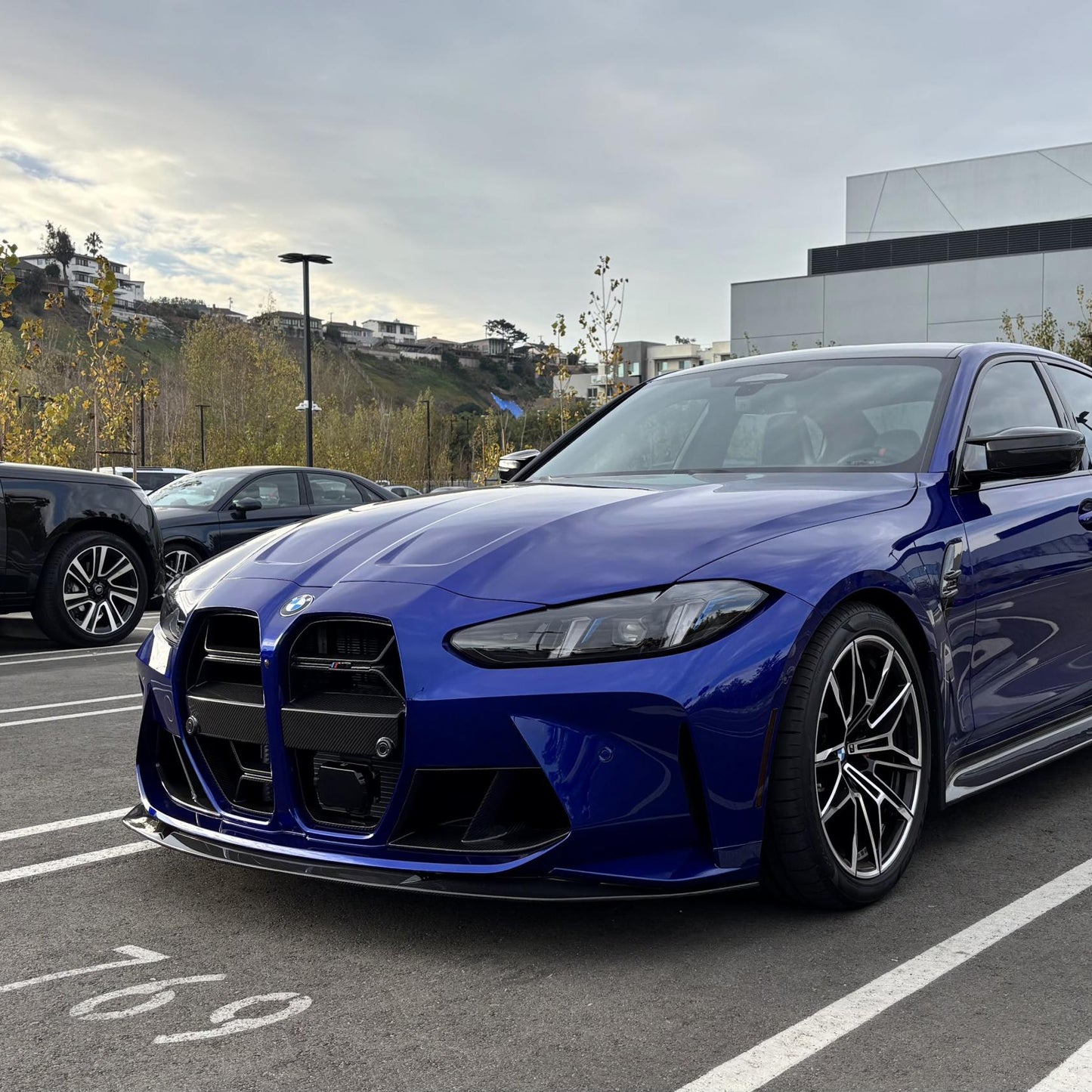 A blue BMW G8X M3/M4 with a carbon fiber front lip installed on the front bumper.